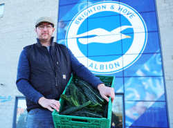Image of Farmer Tom showing off case of vegetables from his farm in front of the American Express Stadium 